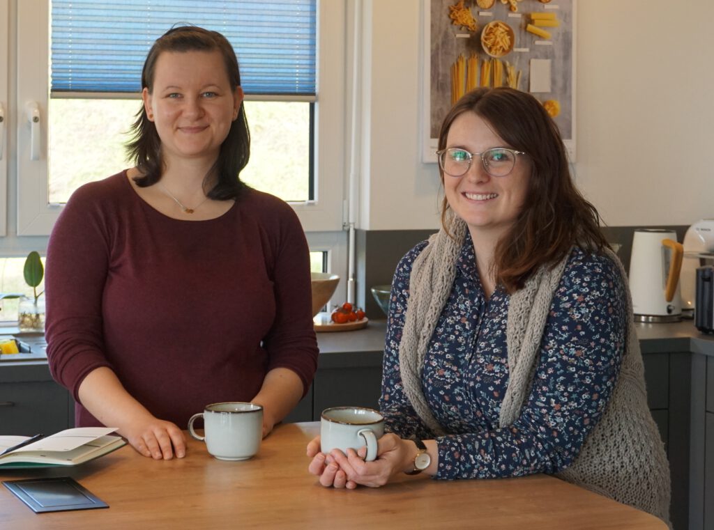 Julia und Jessika stehen in einer modernen Küche hinter dem Tresen und planen bei einer Tasse Kaffee die nächste Veranstaltung für Thüringen bloggt